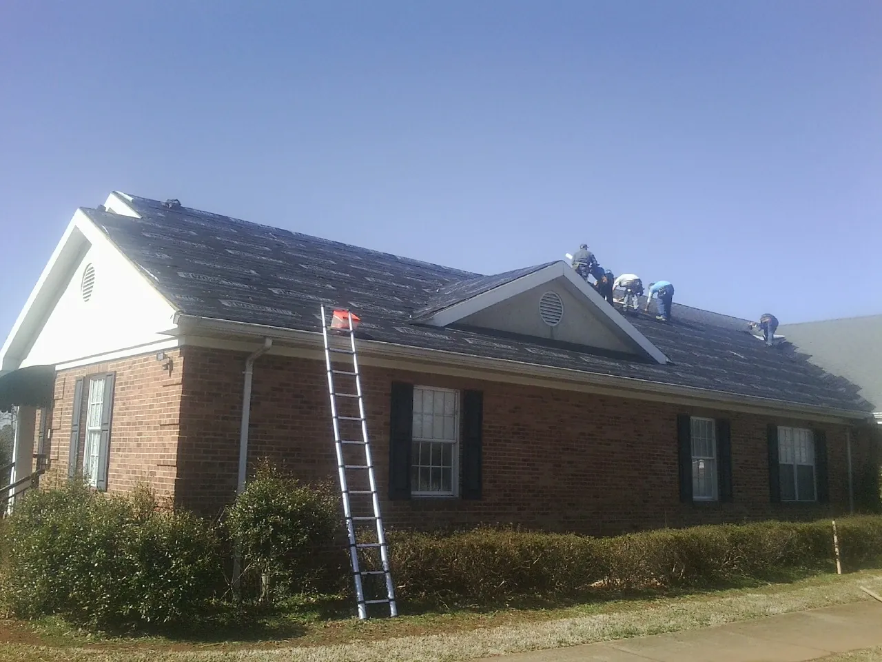Crew tearing shingles off a brick ranch roof with a ladder set — Emergency Roof Repair work by our crew in Belle Mead