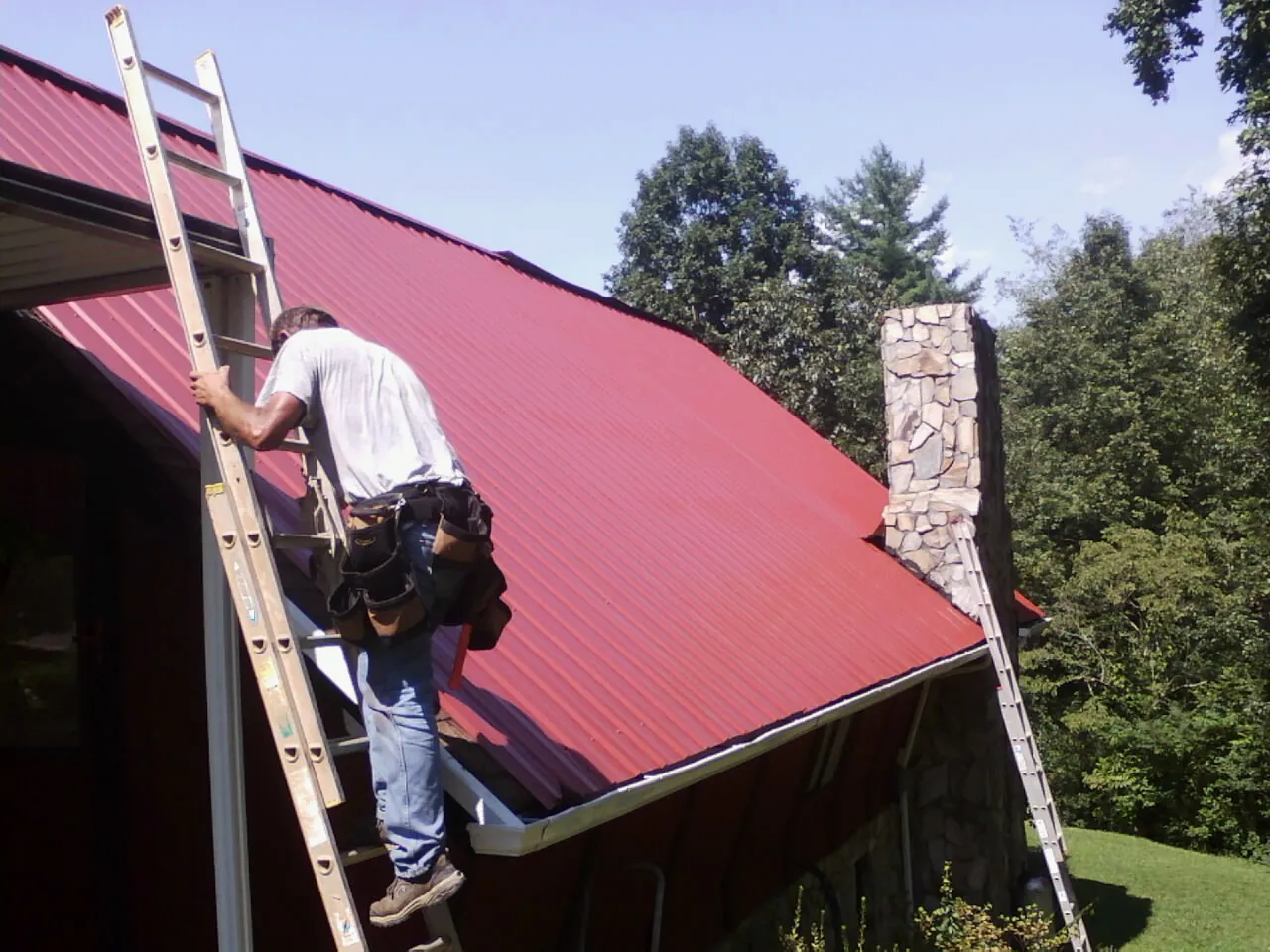 Roofer on a ladder beside a red metal roof and stone chimney — Soffit & Fascia Repair by Roofing Brothers in Belle Mead
